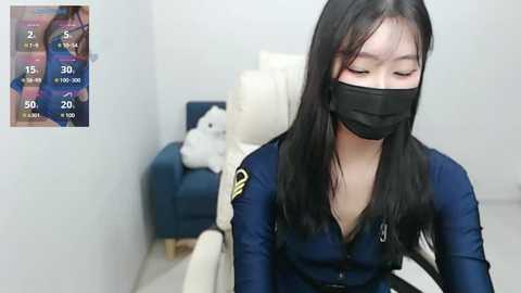 Media: A video of an Asian woman with long black hair and fair skin wearing a navy-blue police uniform and a black face mask, sitting on a white chair in a minimalist room.