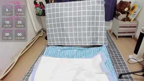 Media: Video of a neatly made bed with a blue and white checkered duvet and a white pillow, surrounded by a gray tiled floor, a radiator, and a teddy bear on a shelf in a cozy bedroom.