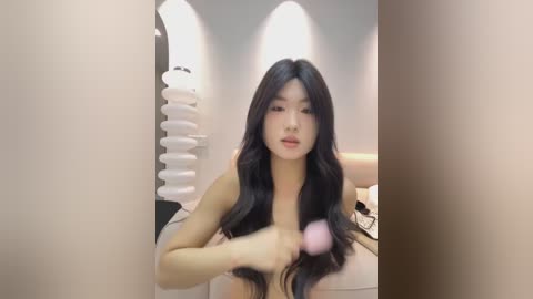 Media: A video of a young Asian woman with long, straight black hair, fair skin, and a slender physique, applying makeup with a pink brush in a modern, minimalist bathroom with white walls and a spiral-shaped towel rack.