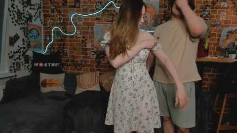 Media: Video of a young woman in a floral dress and a man in a beige shirt and green shorts in a cozy, dimly lit room with a brick wall, posters, and a couch.