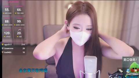 Media: A video of an East Asian woman in a black halter top, wearing a white face mask, adjusting her earbuds, taken indoors with a blurred background.