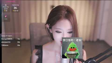 Media: A video of an East Asian woman with straight brown hair, wearing a black top, holding a phone with a green duck emoji, in a blurred hotel room.