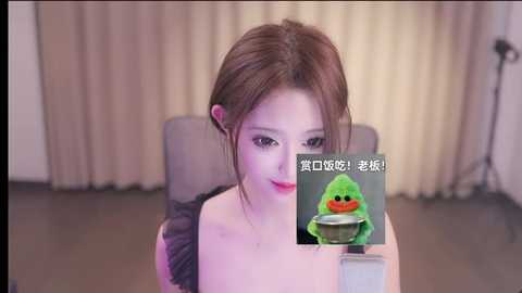 Media: Video of a young woman with fair skin and brown hair, sitting on a gray chair in a beige room. She holds a green plush frog puppet with red eyes. Text overlay in Chinese.