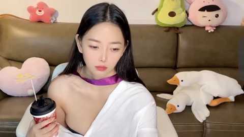 Media: Video of a young Asian woman with long black hair, wearing a white shirt, sipping from a red cup, surrounded by plush toys on a brown couch.