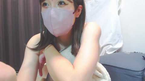 Media: Video of an Asian woman with fair skin, wearing a pink face mask and a sleeveless white top, sitting on a bed with a beige blanket and a gray pillow.