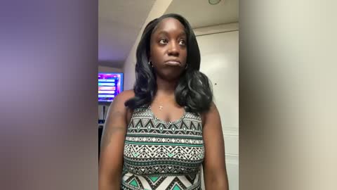 Media: Video of a young Black woman with straight black hair, wearing a geometric-patterned sleeveless top, standing in a brightly lit room with a TV displaying a news channel in the background.