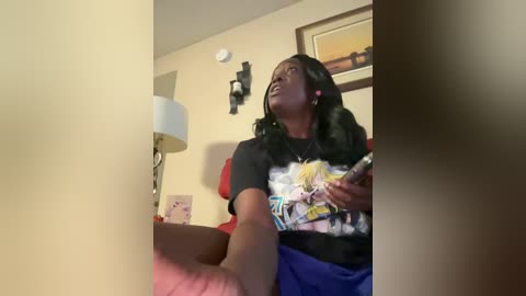 Media: Video of a Black woman with long, wavy hair, wearing a black graphic t-shirt and blue shorts, seated on a red couch, holding a remote, in a beige room with a wall-mounted lamp and framed artwork.