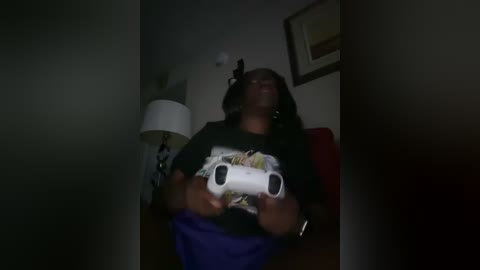 Media: A dimly lit video of a person wearing a black T-shirt with a white video game controller, sitting on a red couch.