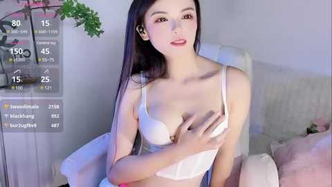 Media: Video of an East Asian woman with fair skin, long dark hair, wearing white lingerie, sitting on a white couch, holding her breast, in a softly lit room.