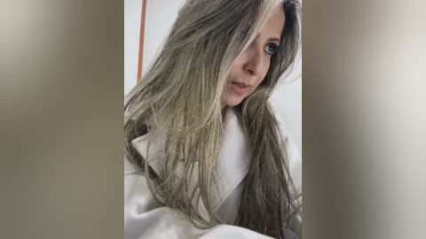 Media: A video of a woman with long, wavy, light gray hair, wearing a white bathrobe, standing in a bathroom. The background is blurred.