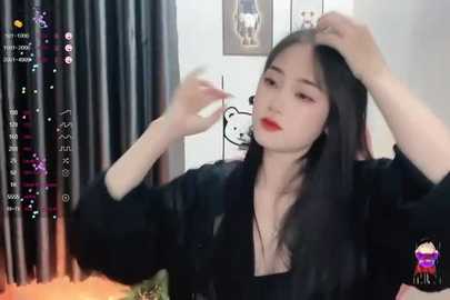 Media: Video of an East Asian woman with long black hair, wearing a black top and red lipstick, fixing her hair in a dimly lit room with a pink pillow and a cartoon character on the wall.