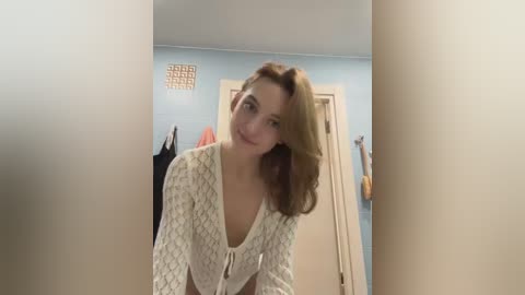 Media: Video of a young woman with shoulder-length wavy brown hair, wearing a patterned white top, smiling in a blue-walled bathroom with towels hanging and a vent on the wall.