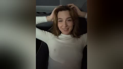 Media: Video of a smiling Caucasian woman with shoulder-length brown hair, wearing a white long-sleeve top, seated in a car, hands in hair, with blurred, out-of-focus background.