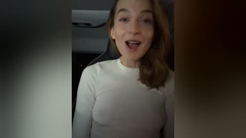 Media: Video of a Caucasian woman with shoulder-length brown hair, wearing a white long-sleeve top, smiling, in a car with grey seats and a dark ceiling.