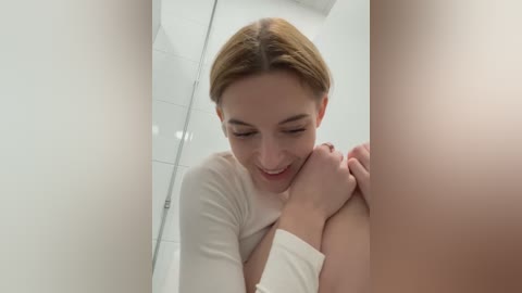 Media: A video of a fair-skinned, blonde woman with a slim build, wearing a white top, smiling while hugging herself in a white-tiled bathroom.