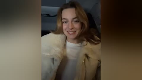 Media: Video of a smiling woman with long, wavy blonde hair, wearing a beige fur coat and white top, seated in a dimly lit car.