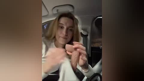 Media: Video of a blonde woman with a neutral expression, wearing a white top, holding a small, white object in her right hand, seated in a car.