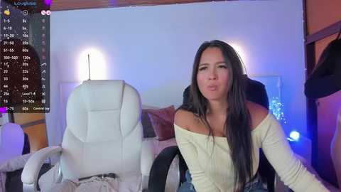 Media: Video of a young woman with long black hair, wearing a white off-shoulder top and blue jeans, sitting on a white gaming chair in a dimly lit room with a calendar and gaming monitor in the background.