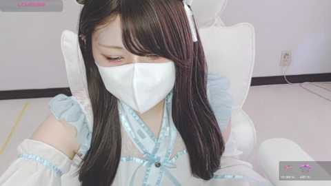 Media: Video of a young Asian woman with long black hair, wearing a white face mask, light blue dress, and sitting in a white chair in a modern room.