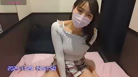 Media: Video of an Asian woman with long black hair, wearing a white off-shoulder top, plaid skirt, and face mask, seated on pink fabric in a modern room with dark walls.