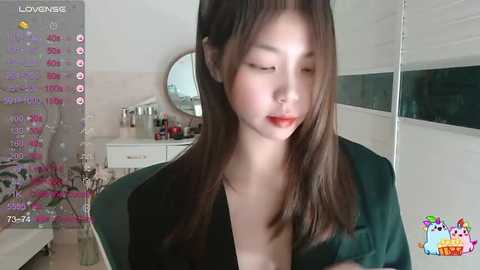 Media: A video of a young Asian woman with fair skin, long brown hair, and red lipstick, wearing a green top, sitting in a dimly lit room with a mirror, pink wall, and a colorful cartoon logo in the bottom right corner.