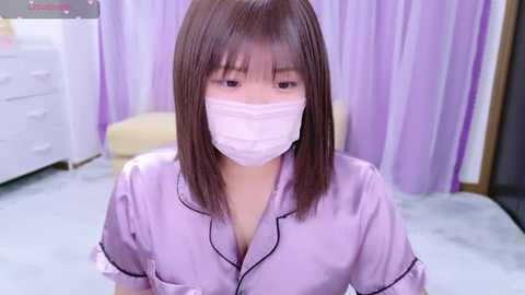 Media: Video of an Asian woman with straight, shoulder-length brown hair, wearing a pastel lavender satin robe with black trim and a white medical mask, sitting on a light carpet in a softly lit room with purple curtains and white dresser.