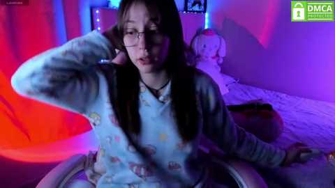 Media: Video of a young woman with long brown hair, glasses, and a light blue floral pajama top, leaning on a bed in a dimly lit bedroom with red and purple lights, a teddy bear, and a pillow in the background.