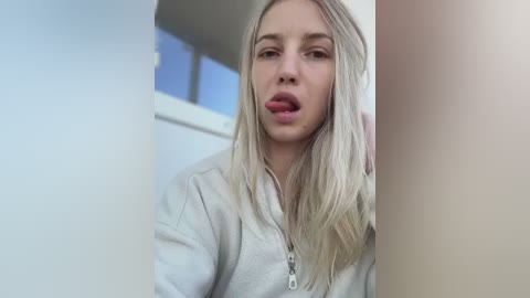 Media: A video of a blonde woman with fair skin and long hair, wearing a white zip-up hoodie, sticking her tongue out while indoors near a window.