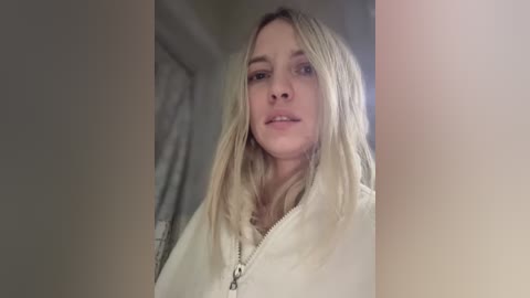 Media: A video of a young woman with long, blonde hair, fair skin, and a casual white hoodie, standing indoors in a dimly lit room with beige walls and a partially visible door.