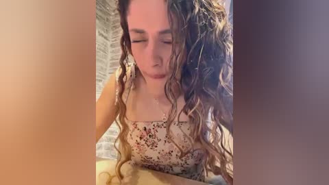 Media: Video of a young woman with long, wet, curly brown hair, wearing a floral dress, sitting on a bed with a beige blanket, indoors with a brick wall background.