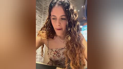 Media: Video of a young woman with long, curly, light brown hair, wearing a floral dress, sitting at a table in a cozy, brick-walled caf\u00e9.