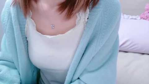 Media: Video of a fair-skinned woman with shoulder-length auburn hair, wearing a white lace-trimmed camisole under a light blue fleece robe, with a delicate necklace. Background includes a bed with light-colored pillows.