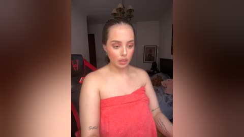 Media: A video of a young, light-skinned woman with a slender build, wearing a red towel, standing in a dimly lit bedroom. Her hair is tied up in a bun.