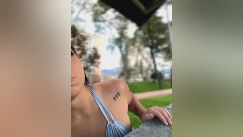 Media: A video of a young woman with light skin and a tattoo reading \"XXX\" on her left shoulder, wearing a light blue halter top, lying on a stone bench in a grassy park.