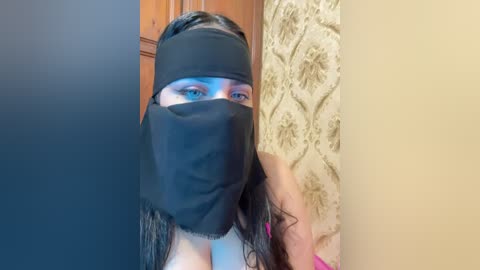 Media: A video of a woman with long black hair and fair skin, wearing a black mask, blue eyeshadow, and pink nails, standing in a room with floral wallpaper and wooden furniture.