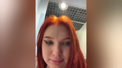 Media: Video of a young woman with striking, long, bright red hair and heavy black winged eyeliner, smiling slightly. Background shows a modern, tiled ceiling with a recessed light.