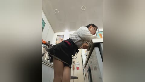 Media: A video of a slender East Asian woman in a kitchen, wearing a gray sweater, black apron, and black skirt, bent over a stove, with a blurred background and white walls.