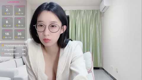 Media: A video of an East Asian woman with straight black hair, wearing glasses, a white blazer, and sitting in a modern, minimalist living room with white walls, green curtains, and a beige sofa.