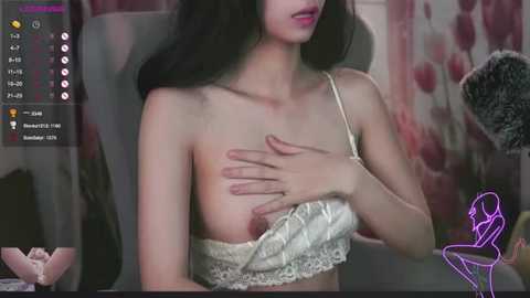 Media: A video of a young East Asian woman with fair skin and long black hair, wearing a lacy white bra, covering her breast with her hand, in a dimly lit room with floral wallpaper.