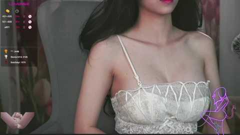 Media: Video of an East Asian woman with long black hair, wearing a lace white bralette, standing against a gray background.