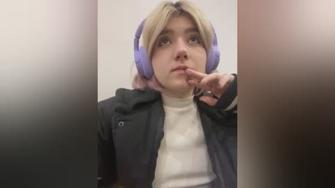 Media: Video of a young woman with light skin, blonde hair, and purple headphones, wearing a black jacket over a white turtleneck, with her finger to her lips, against a blurred background.