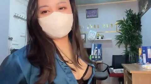 Media: Video of an Asian woman with long, dark hair wearing a white surgical mask, blue scrubs, and a black necklace. Background shows a desk with a computer, a potted plant, and a wall with a sign.