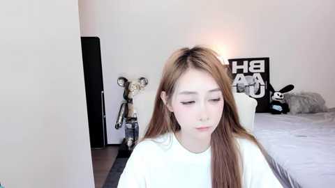 Media: Video of a young Asian woman with long, straight brown hair, wearing a white t-shirt, sitting on a white chair in a minimalist bedroom with a plush toy rabbit and a black \"HEART\" poster on the wall.