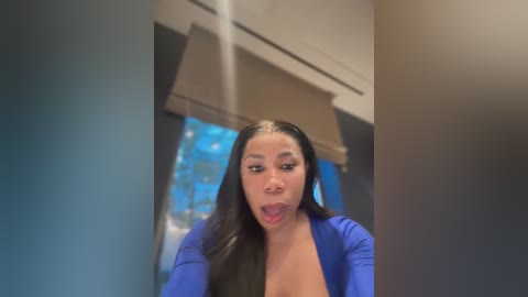 Media: Video of a young Black woman with long straight hair, wearing a blue top, and looking surprised, in a modern kitchen with a blue-tiled backsplash and beige window shade.