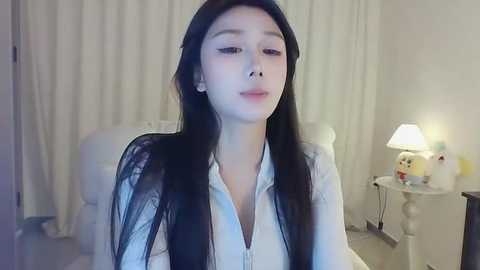 Media: Video of a young East Asian woman with long black hair, pale skin, and a slender physique, wearing a white button-down shirt, sitting in a softly lit room with white curtains, a white chair, and a lamp.