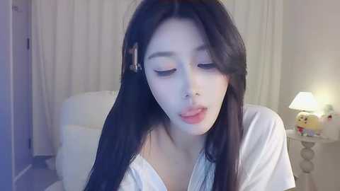 Media: Video of a young East Asian woman with long, straight black hair, wearing a white shirt, sitting indoors, with a white lamp, plush toys, and white curtains in the background.