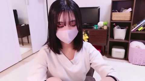 Media: Video of an Asian woman with straight black hair and a white mask, sitting at a desk in a tidy room with a TV, shelves, and laundry basket.