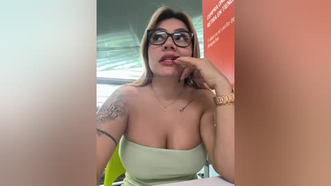 Media: Video of a curvy, light-skinned woman with long blonde hair, wearing glasses, a strapless light green dress, and a gold watch. She has tattoos on her left arm. Background shows blurred office setting with a red and white poster.