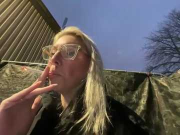 Media: Video of a blonde woman with glasses, looking contemplative, standing against a corrugated metal wall and camouflage tarp in an outdoor setting with a clear blue sky.