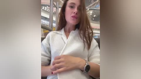 Media: A video of a young Caucasian woman with long brown hair, wearing a white ribbed shirt and silver watch, sitting in a modern, brightly lit indoor setting with glass railings and visible ceiling.